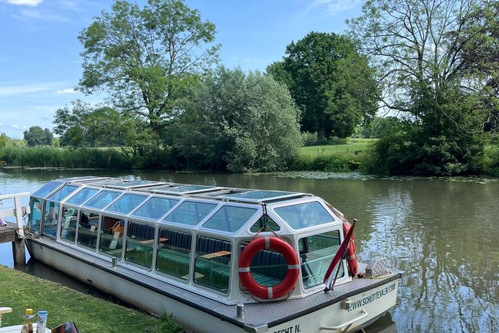 Glass-roofed boat docked on a river beside a grassy bank with trees.