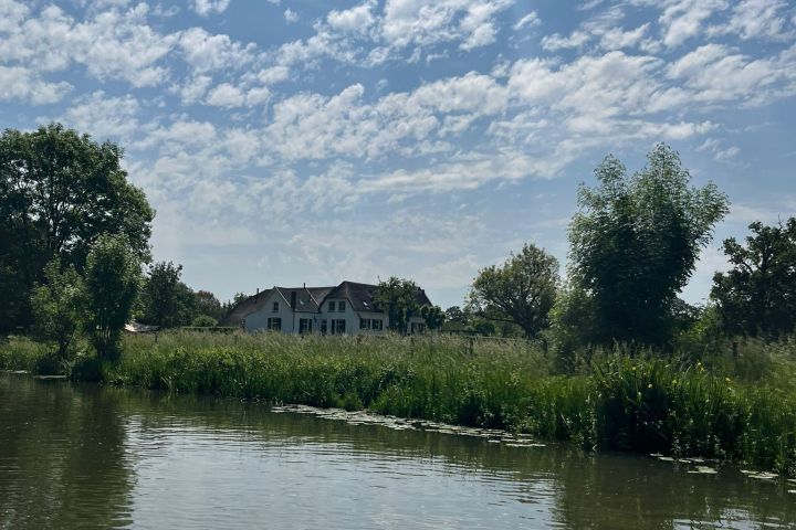 House by a tree-lined river under a partly cloudy sky.