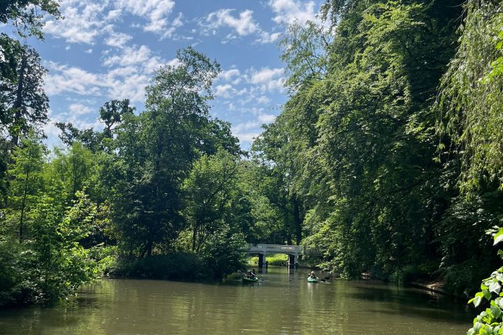 Lush trees surround a calm river with a small bridge in the distance under a blue sky with clouds.