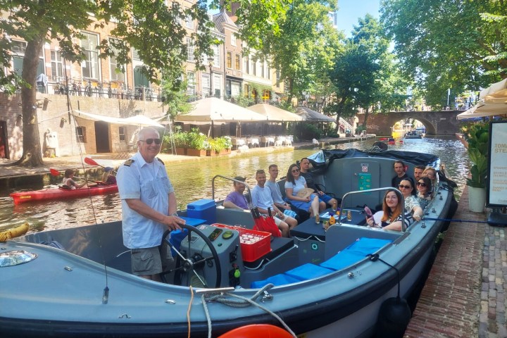 Group of people on a canal boat with trees and cafes nearby.