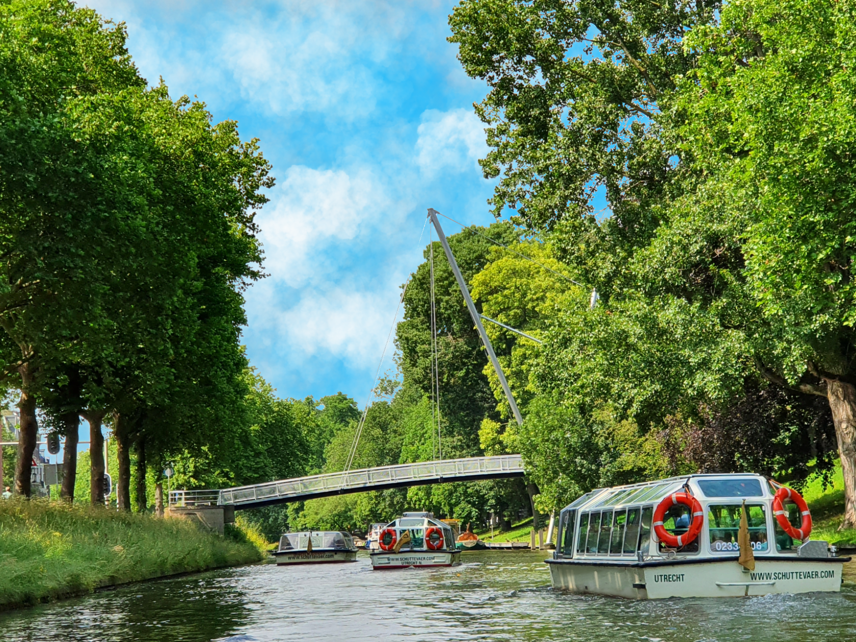 Boats on a tree-lined canal under a bridge with a blue sky in the background.