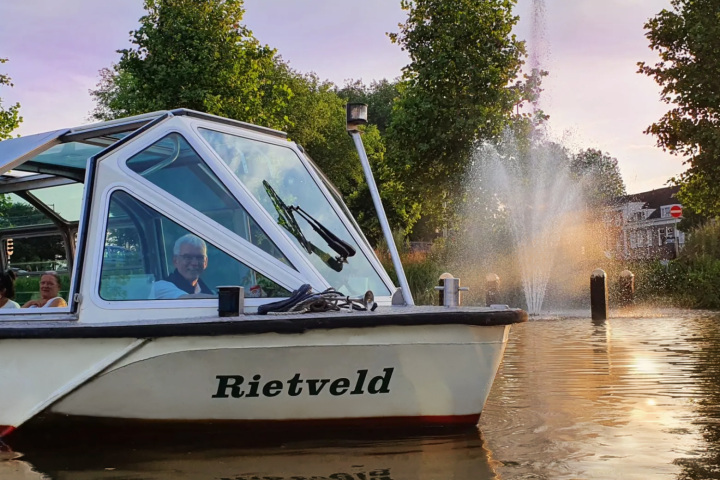 White boat named Rietveld on water with fountain and trees in background during sunset.