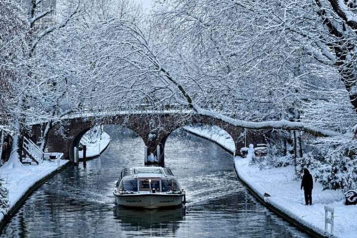 Boat on snowy canal under bridge with snow-covered trees.