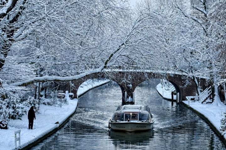 Boat on snowy canal under bridge with snow-covered trees.
