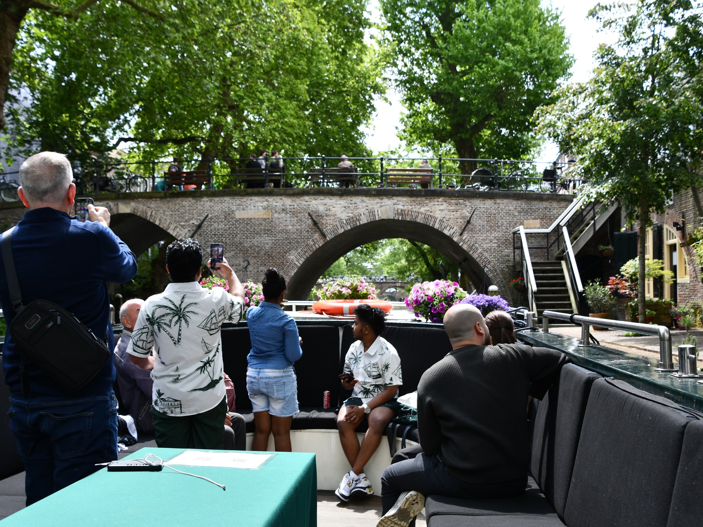 People on a boat tour photographing a scenic stone bridge surrounded by trees.