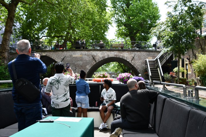 People on a boat tour photographing a scenic stone bridge surrounded by trees.