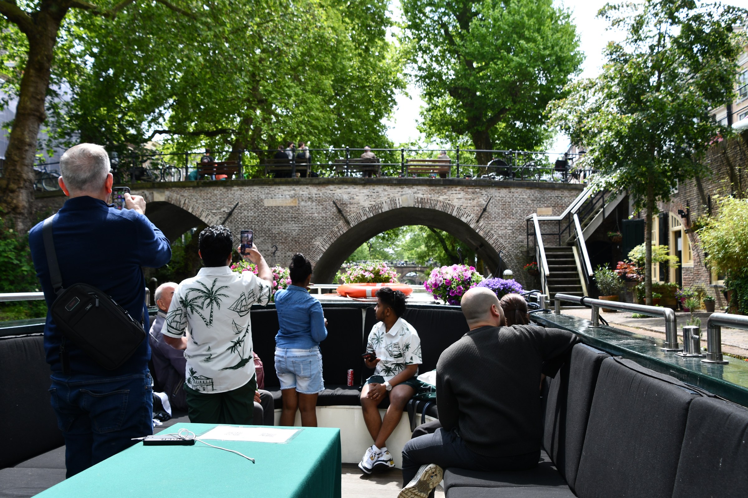 People on a boat tour photographing a scenic stone bridge surrounded by trees.