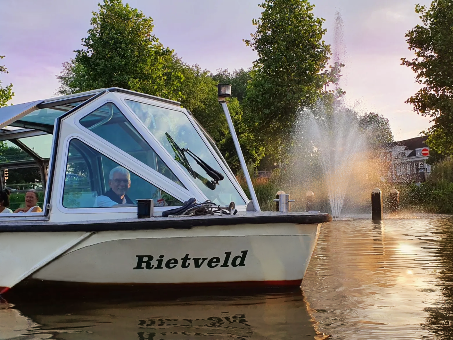 White boat named 'Rietveld' on water near a fountain and trees with people inside during sunset.