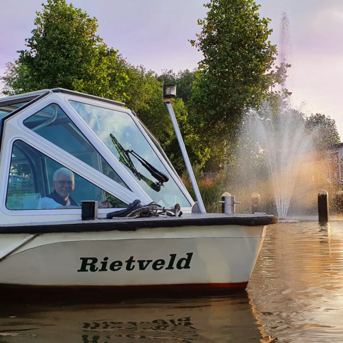 White boat named 'Rietveld' on water near a fountain and trees with people inside during sunset.