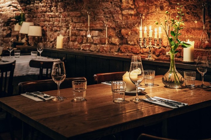 Cozy restaurant with a wooden table, wine glasses, candles, and flowers against a brick wall.