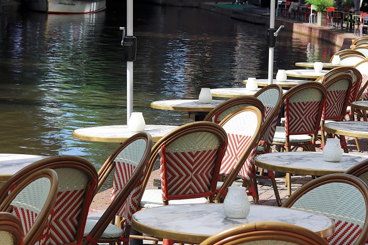 Outdoor café with empty tables and chairs by a canal with a boat and bridge.