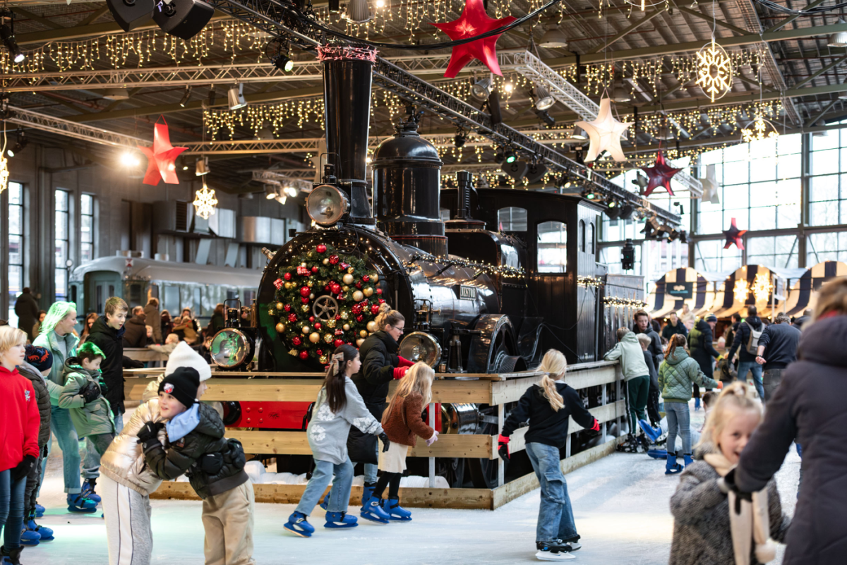 Kids ice skating around a train in a festive indoor setting with lights and decorations.