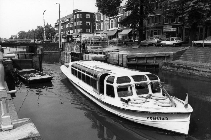 Black and white photo of a canal boat named Domstad passing through a lock with buildings in the background.