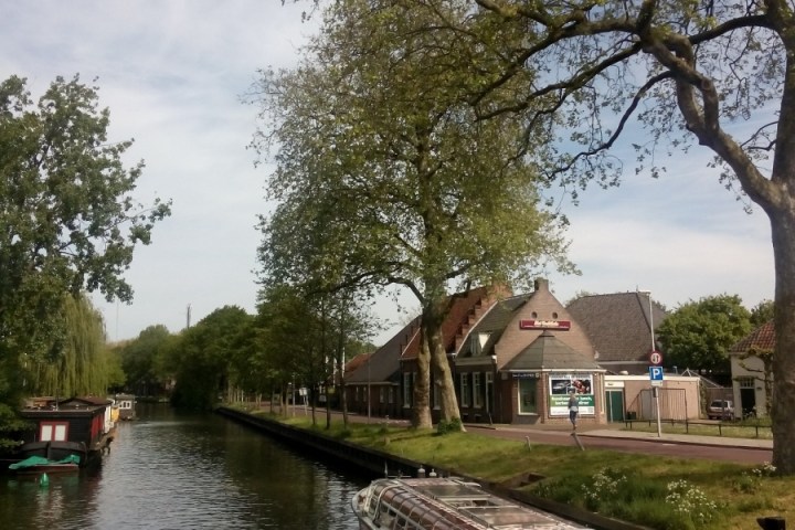Canal with a glass-roofed boat, trees, and a brick building under a partly cloudy sky.