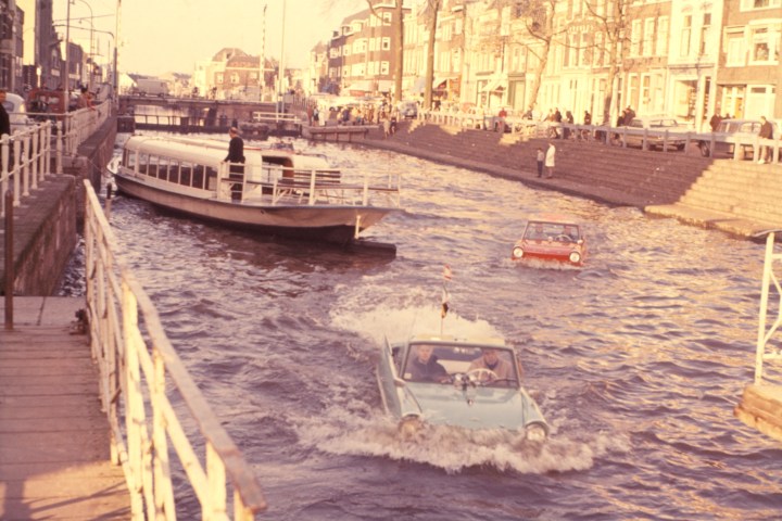 Amphibious cars in a canal with a boat, buildings, and stairs along the water in a cityscape.