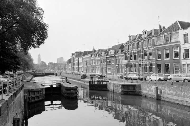 Black-and-white photo of a canal with bridge, cars, and historic buildings alongside in a city.