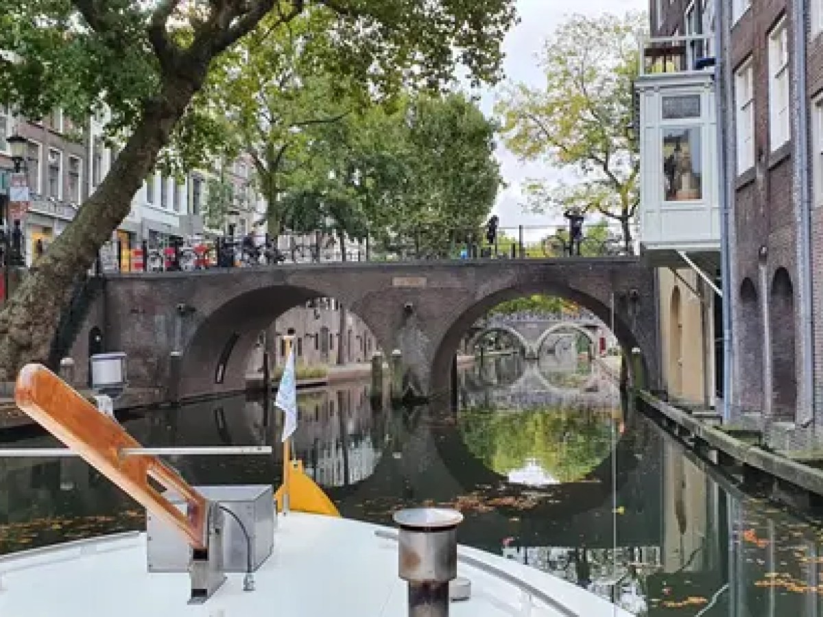 Boat on a canal under a bridge in a city with trees and buildings.