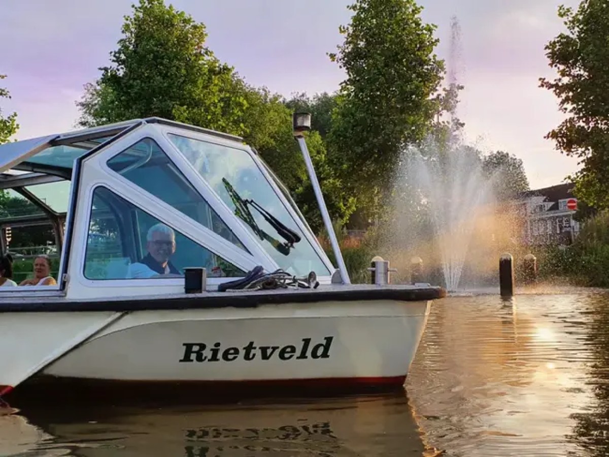 Boat named Rietveld on water near fountain and trees, with people inside.