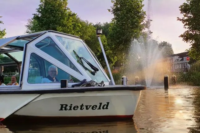 Boat named Rietveld on water near fountain and trees, with people inside.