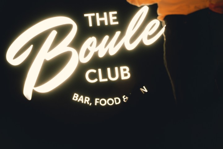 Person in white shirt standing near illuminated 'The Boule Club' sign.