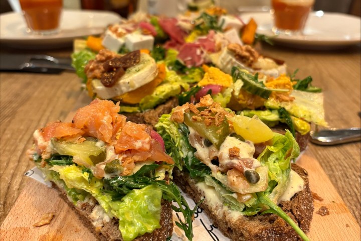 Assorted open sandwiches with toppings on a wooden board, with drinks in the background.