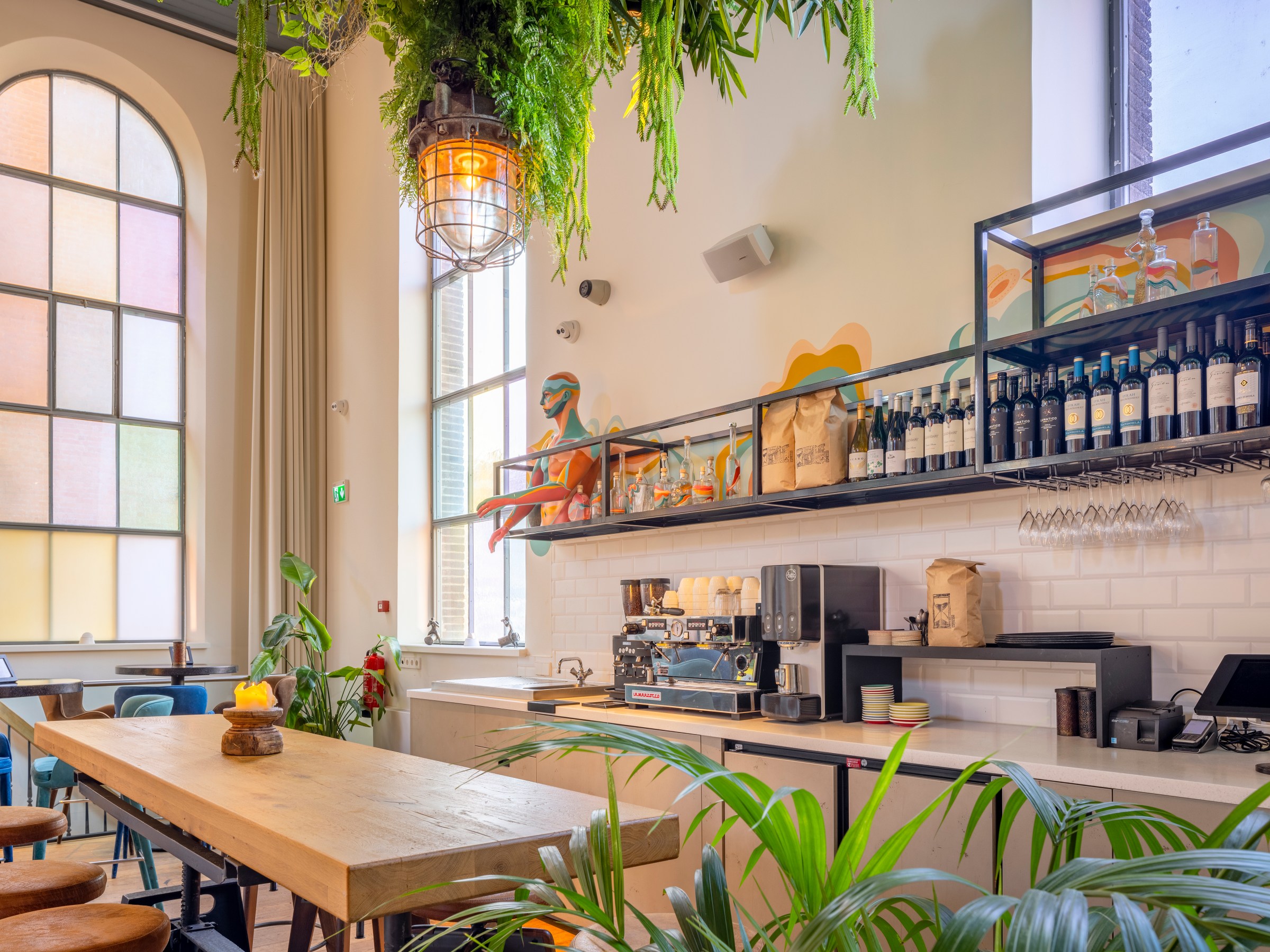 Modern cafe with wooden tables, hanging plants, and a coffee machine on a white tiled counter.