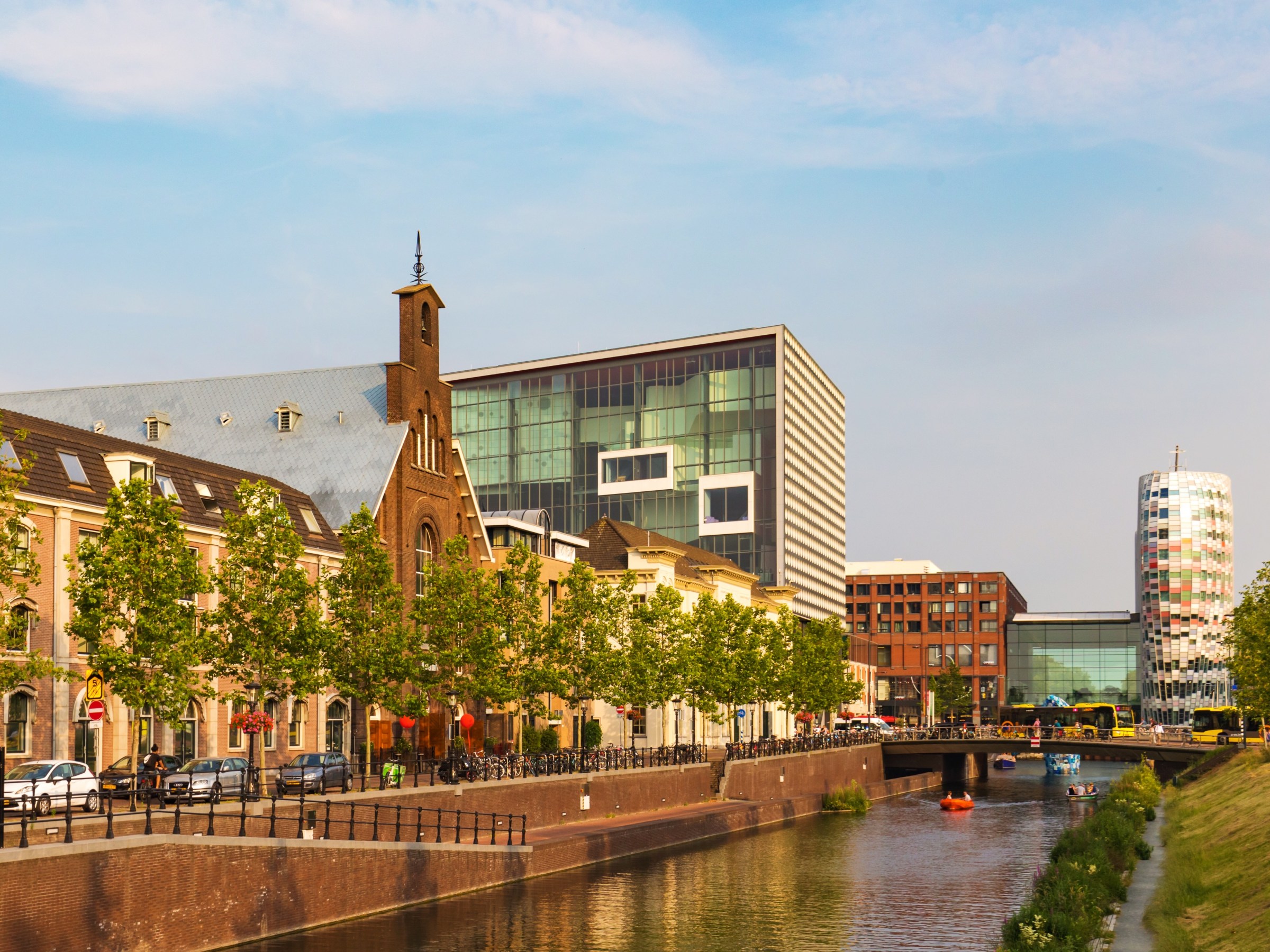 Canal with modern and historic buildings, trees, and a bridge under a blue sky.