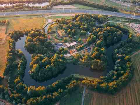 Aerial view of a lush green island surrounded by a river, with buildings and trees, near farmlands and a road.