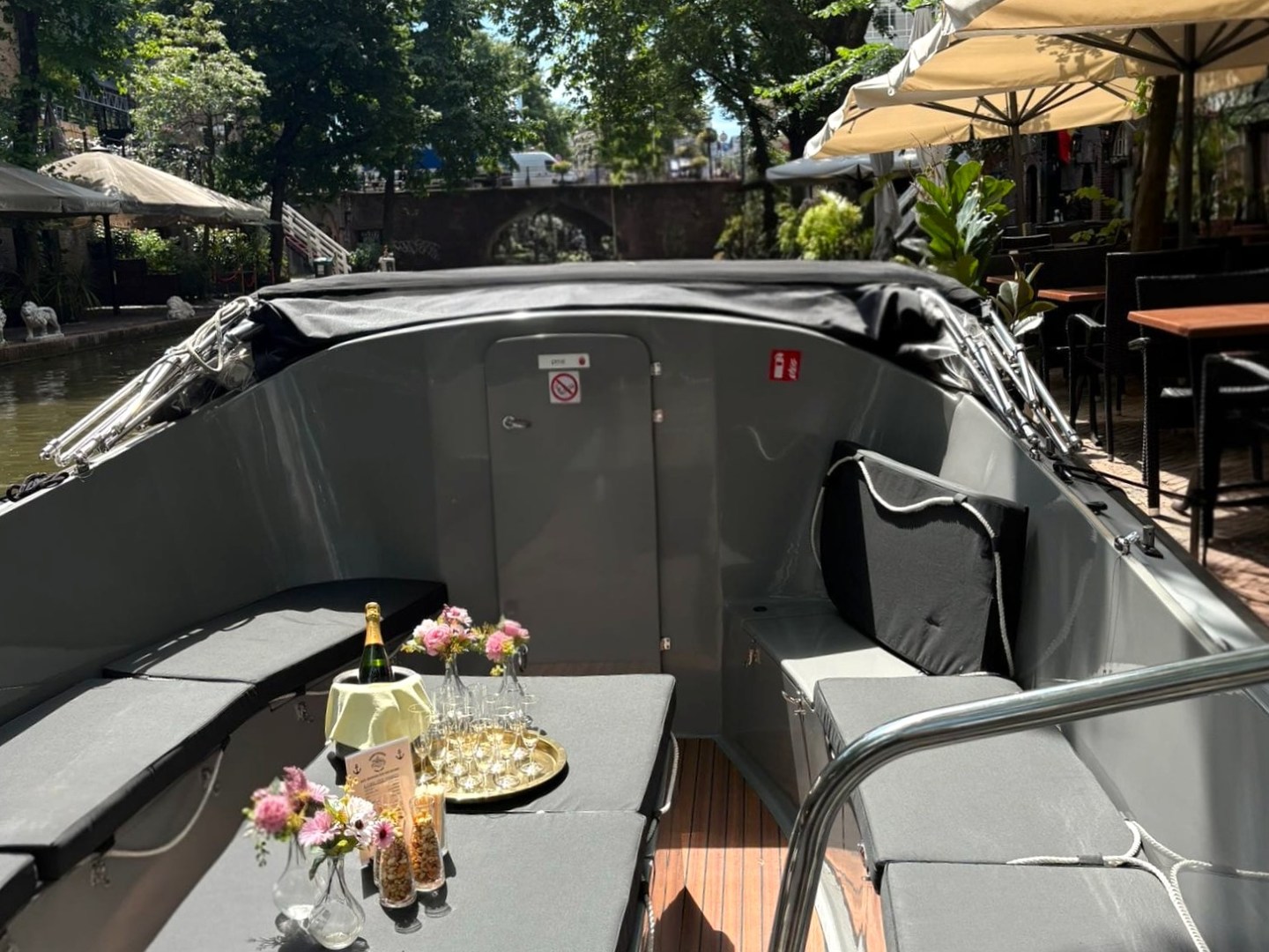 Cozy boat seating with flowers and drinks under sunny trees.