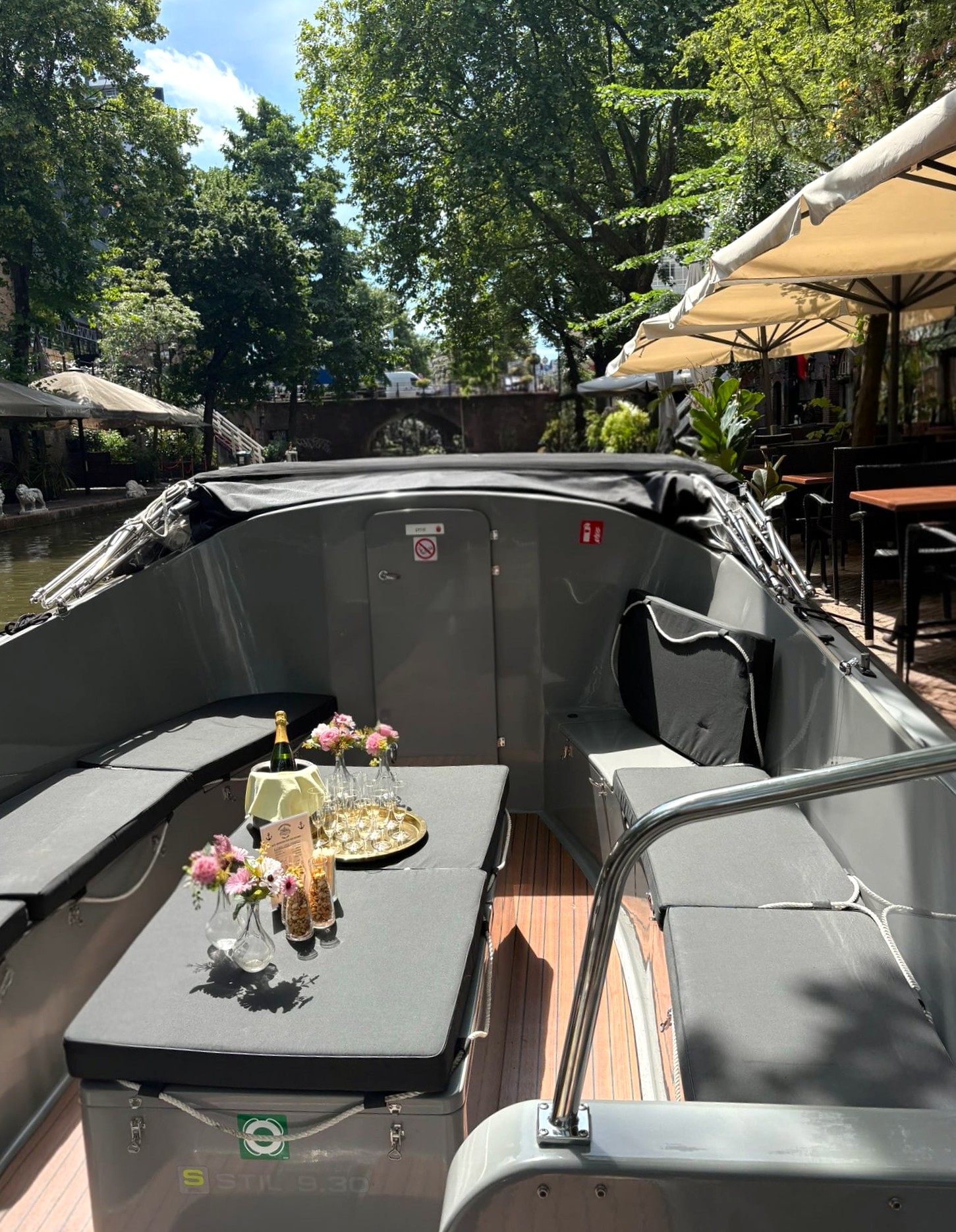Cozy boat seating with flowers and drinks under sunny trees.