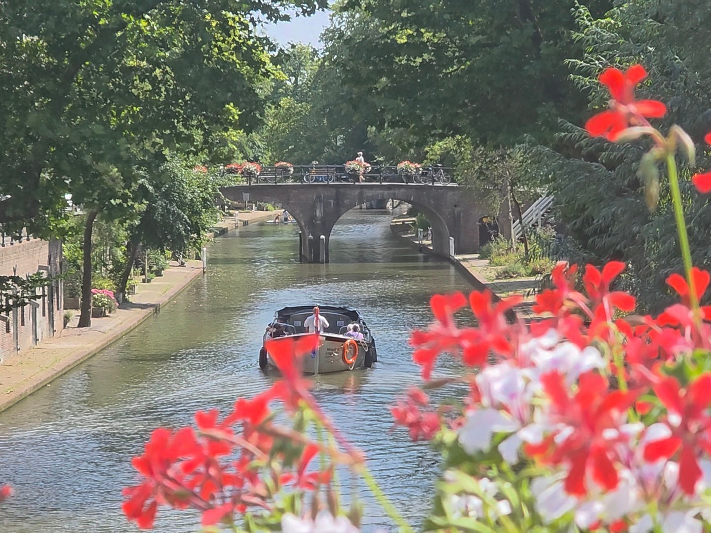 Boat on canal with bridge and red flowers in foreground.