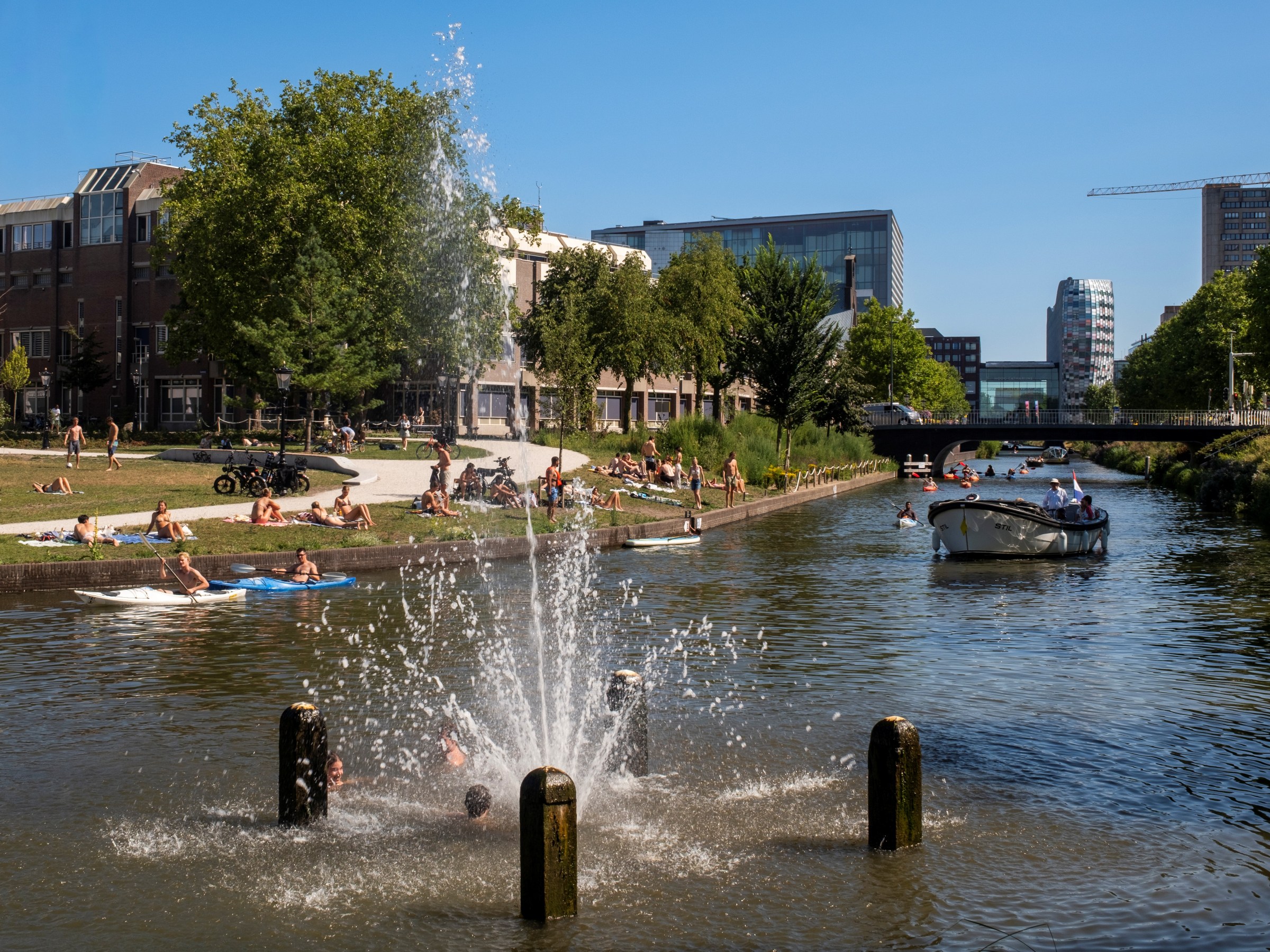 People relaxing by a canal with a fountain and boats on a sunny day.