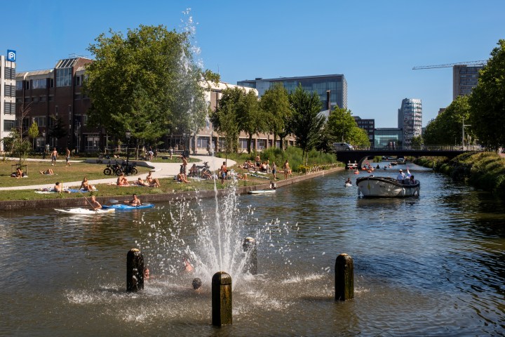 People relaxing by a canal with a fountain and boats on a sunny day.