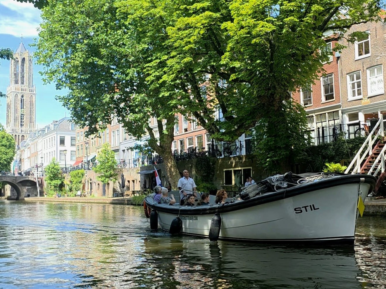 Boat on canal with trees, buildings, and a church tower in the background.
