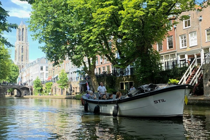 Boat on canal with trees, buildings, and a church tower in the background.